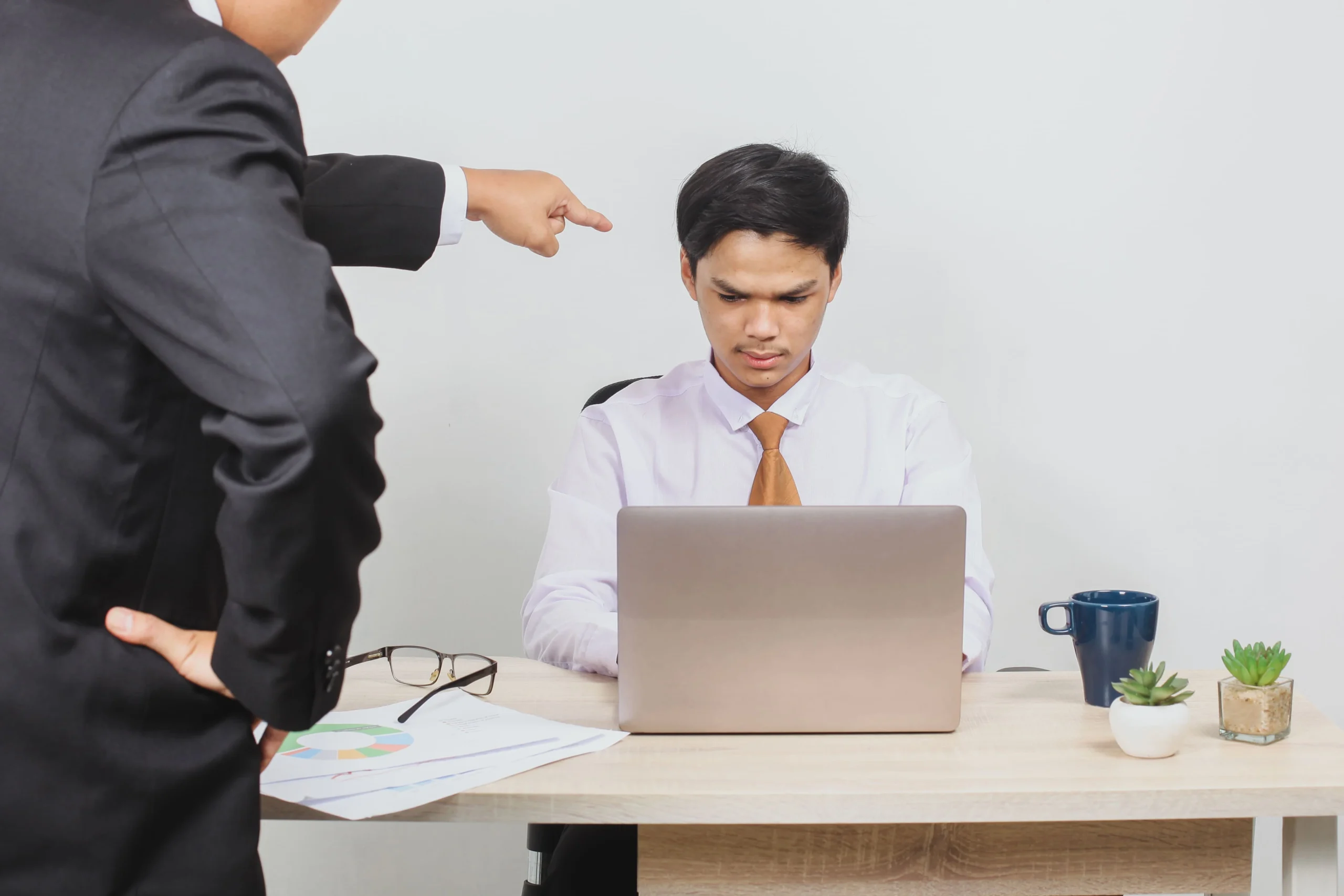 an image representing the blog An employee sits at a desk looking stressed while working on a laptop, as a manager in a suit points sternly at him, highlighting workplace conflict.