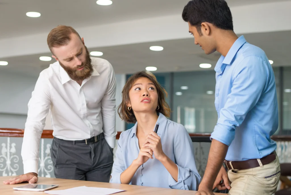 A female employee seated at an office desk looking concerned while speaking with two male colleagues leaning over her table, illustrating workplace disputes and discrimination claims under California FEHA laws.