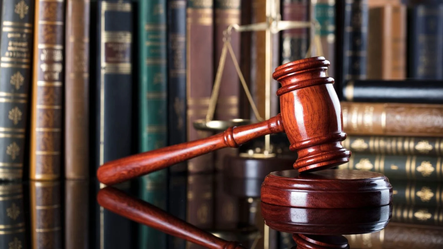 Wooden gavel resting on a sound block with legal books and justice scales in the background, representing labor laws on required hours between shifts.