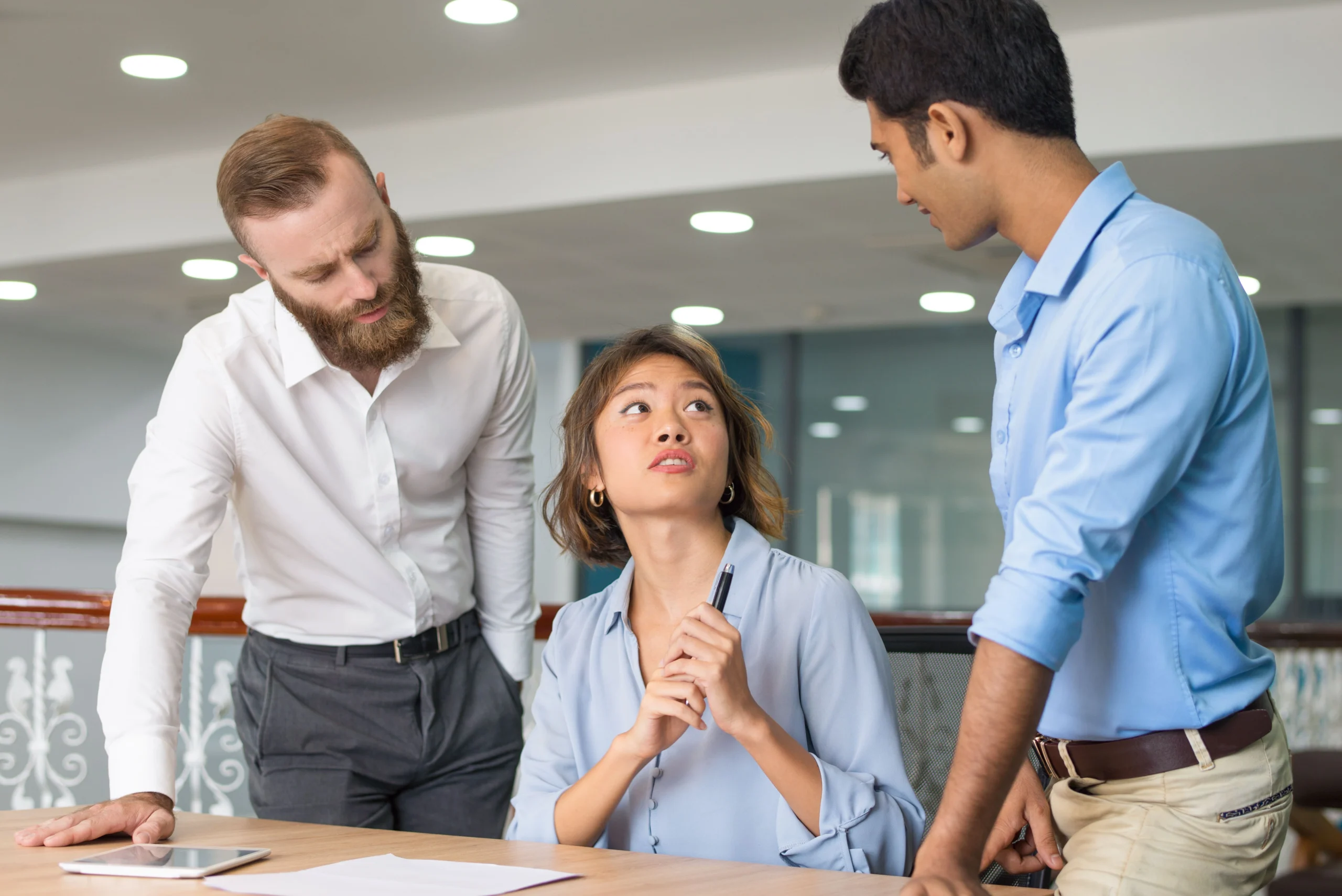 A female employee seated at an office desk looking concerned while speaking with two male colleagues leaning over her table, illustrating workplace disputes and discrimination claims under California FEHA laws.